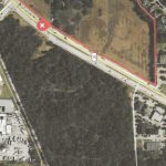 Aerial view of a plot of land outlined in red, bordered by roads and surrounded by buildings, trees, and parking lots. A red "X" marks the entrance on a main road passing alongside the plot.