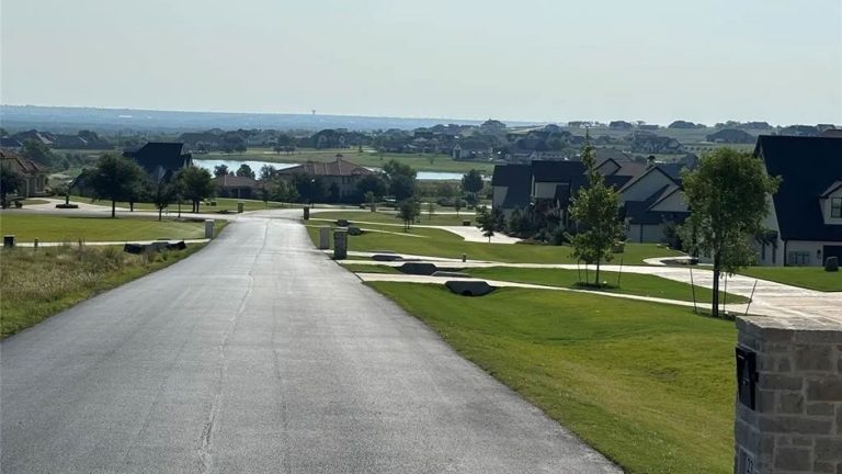 A paved road winds through the quiet Aledo neighborhood, 76008, with well-kept lawns, modern houses, and trees, overlooking a distant lake under a hazy sky.