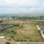 Aerial view of a large, mostly empty plot of land outlined in red, surrounded by suburban homes, roads, and greenery under a partly cloudy sky.