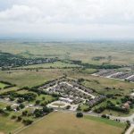 Aerial view of a suburban neighborhood with clusters of houses, roads, and green open fields stretching into the distance under a cloudy sky.