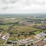 Aerial view of a suburban area with roads, buildings, houses, and open green fields under a cloudy sky, stretching out to the horizon.