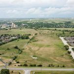 Aerial view of a suburban area with clusters of houses surrounding a large open grassy field, sparse trees, and roads under a partly cloudy sky.