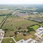 Aerial view of a semi-rural area with open fields, scattered trees, residential neighborhoods, and commercial buildings, intersected by roads and a railway line under a cloudy sky.