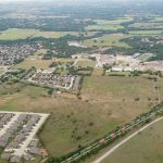 Aerial view of a suburban area with clusters of houses, open fields, a road with a train, and scattered trees, with more residential neighborhoods and green spaces in the background.