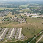 Aerial view of a suburban area with rows of houses, open green fields, a railroad track, and clusters of trees scattered throughout the landscape under a hazy sky.