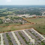 Aerial view of a suburban area with a large, irregularly shaped open field outlined in red, surrounded by houses, roads, and scattered trees under a partly cloudy sky.