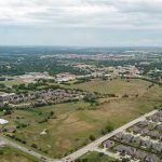 Aerial view of a suburban area with rows of houses, open green fields, scattered trees, and roads under a cloudy sky. In the distance, more buildings and a small town are visible.