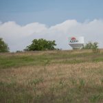 A white water tower labeled "Aledo" stands behind a grassy, slightly hilly field with scattered trees under a blue sky with white clouds.