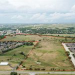 Aerial view of a large open grassy field bordered by suburban houses on three sides, with the property boundary marked by a red outline. Rolling hills and a distant town are visible under a cloudy sky.