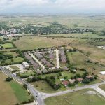 Aerial view of a suburban neighborhood with houses, streets, and green fields surrounding the area. Roads and scattered buildings are visible, with open countryside stretching into the distance under a cloudy sky.