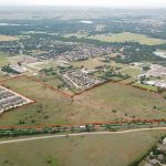 Aerial view of a suburban area with a large open field outlined in red, surrounded by residential neighborhoods, roads, and patches of trees. A train is visible along the bottom edge of the outlined area.