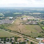 Aerial view of a suburban area with clusters of houses, open green fields, roads, and tree-lined neighborhoods. The landscape extends to the horizon under a cloudy sky.