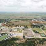 Aerial view of a rural area with buildings, a football field, roads, and an open field bordered in red, surrounded by trees and open land under a cloudy sky.