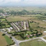 Aerial view of a suburban residential area with rows of houses, surrounding green fields, roads, and scattered buildings under a cloudy sky. Spacious open land and neighborhoods are visible in the background.