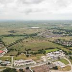 Aerial view of a semi-rural area with clusters of buildings, roads, and houses surrounded by open green fields and patches of trees under a cloudy sky.