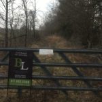 A metal gate blocks a narrow, overgrown path lined with leafless trees. The gate has two signs, one advertising a land business with contact info, and another smaller, unreadable sign. Fields and cloudy sky are visible in the background.