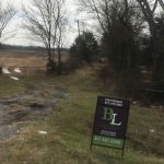A rural dirt road leads to a gated field with trees and overcast sky. A real estate sign for "BL" is posted in the grass by the road with contact information for Carl Roland.