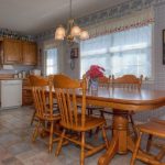 A cozy kitchen and dining area with wooden cabinets, a large wooden dining table and chairs, tile flooring, and bright windows with white curtains. Shelves above the cabinets hold bottles and decor items.