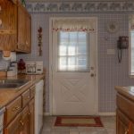 A cozy kitchen with wooden cabinets, a double sink, and small appliances on the counter. A door with a window is centered, flanked by windows with white blinds and valances. The walls have blue plaid wallpaper and floral borders.