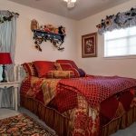A cozy bedroom with a red and gold comforter set, two windows with blue-striped curtains, sunflowers above the windows, teddy bears on a wall shelf, and a small white nightstand with a red lamp.