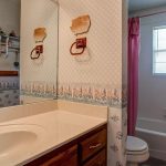 A bathroom with a large mirror, single sink vanity, floral wallpaper, and wooden accents. A bathtub with a pink shower curtain and a window is next to a toilet. The room has vintage decor and patterned walls.