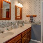 A bathroom with dual sinks, wooden cabinets, two mirrors with wooden frames, patterned wallpaper, and a towel hanging on a wooden rack. There are toiletries on the countertop and an open door leading to another room.
