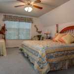 A cozy bedroom with a large bed and a single bed, both with floral bedding. There is a ceiling fan, window with a valance, wooden nightstand, and decorative items on the walls. The room has soft lighting and carpeted floor.