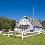 A two-story, light blue farmhouse with a balcony and white fencing sits on a grassy lawn under a clear blue sky, surrounded by trees.