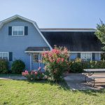 A blue two-story house with white trim, surrounded by green grass, flowering bushes, and trees. A picnic table sits on a concrete patio in front of the house under a clear blue sky.