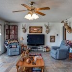 A cozy living room with tiled floor, two blue recliners, a TV on a stand, mounted deer heads on the wall, a ceiling fan with lights, and rustic decor, including a stone fireplace and wood coffee table.