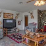 A cozy living room with reclining chairs, a large TV, mounted deer heads, a red wood-burning stove, stone accent wall, and Texas-themed decor. The coffee table holds wood carvings and magazines.
