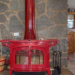 A red wood-burning stove with a chimney pipe stands against a stone wall. A small basket with firewood is on the left. The kitchen with wooden cabinets and a dining area is visible in the background.