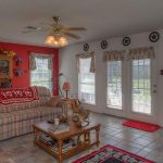 A cozy living room with a plaid sofa, red accent wall, large windows with floral curtains, a ceiling fan, coffee table, and Texas star decorations above the windows. Sunlight streams through the glass doors.