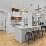 Modern kitchen with white cabinets, a large island with four stools, pendant lights, stainless steel appliances, wood flooring, and an open layout leading to a game area with a pool table and large windows.
