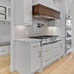 Modern kitchen with white cabinets, gold handles, a large stainless steel stove and oven, a wooden range hood, tile backsplash, light wood floors, and a small built-in bench with shelves in the entryway.