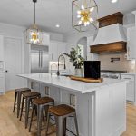 Modern kitchen with white cabinets, a large center island with three wooden stools, stainless steel appliances, gold pendant lights, wood range hood, and light wood flooring. A plant and tray decor are on the island.