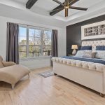 Bright, modern bedroom with large window, beige tufted bed, navy and gray bedding, two wooden nightstands with lamps, a tan chaise lounge, potted plant, and wall art above the bed. Light wooden floor throughout.