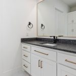 A modern bathroom features white cabinets with gold handles, a black granite countertop, a rectangular sink, a large framed mirror, a towel ring, and a glimpse of a shower with a window in the background.