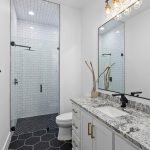 Modern bathroom with a glass-enclosed shower, white subway tiles, black fixtures, hexagonal black floor tiles, a granite countertop, and a large mirror above the vanity with decorative branches in a vase.