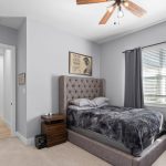 A modern bedroom with gray walls, a tufted bed with gray bedding, two pillows, a wooden nightstand, gray curtains, a ceiling fan, and a window letting in natural light. A hallway is visible through an open door.