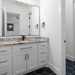 Modern bathroom with white cabinets, black handles, granite countertop, large mirror, black hexagonal floor tiles, and a view of adjacent built-in storage cabinets in a hallway with blue hex tile flooring.