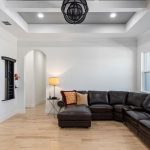 Modern living room with a dark brown leather sectional sofa, accent pillows, a floor lamp, and light wood flooring. An open kitchen area with white cabinets and a wine cooler is visible on the left. The ceiling has recessed lighting.