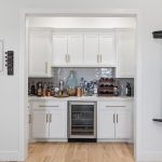 A home bar area with white cabinets, a wine fridge, shelves of bottles, and a dartboard mounted on the left wall. The right wall has a pool cue rack and billiard accessories. Light wood flooring throughout.