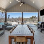 A covered outdoor patio with a long wooden dining table, cushioned seating, a grill, mini fridge, TV, and a pool in the background, overlooking a natural landscape with trees.