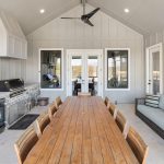 Modern covered patio with long wooden dining table and chairs, built-in stone and stainless steel grill area, flat-screen TV, ceiling fan, and cushioned outdoor sofa; large windows and glass doors provide ample natural light.