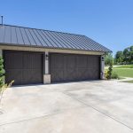 Wide concrete driveway leading to a modern house with three dark wooden garage doors, surrounded by landscaped plants and green lawn under a clear blue sky.