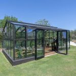 A glass greenhouse with plants inside sits on a well-manicured lawn near a curved concrete pathway, surrounded by green trees and bushes under a clear blue sky.