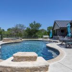 A modern backyard with a kidney-shaped swimming pool, attached hot tub, patio seating, tables with blue umbrellas, and a house with a metal roof, surrounded by greenery under a clear blue sky.