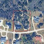 Aerial view of a residential neighborhood with several houses, driveways, and surrounding trees. Two adjacent lots are outlined in red, one mostly wooded and the other containing a house and driveway. Streets curve around the lots.