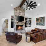 A modern living room with a stone fireplace, wall-mounted TV, brown leather sofas, armchair, metal side table, built-in shelves, large ceiling fan, wooden floors, and white walls. A peacock painting hangs on the right wall.